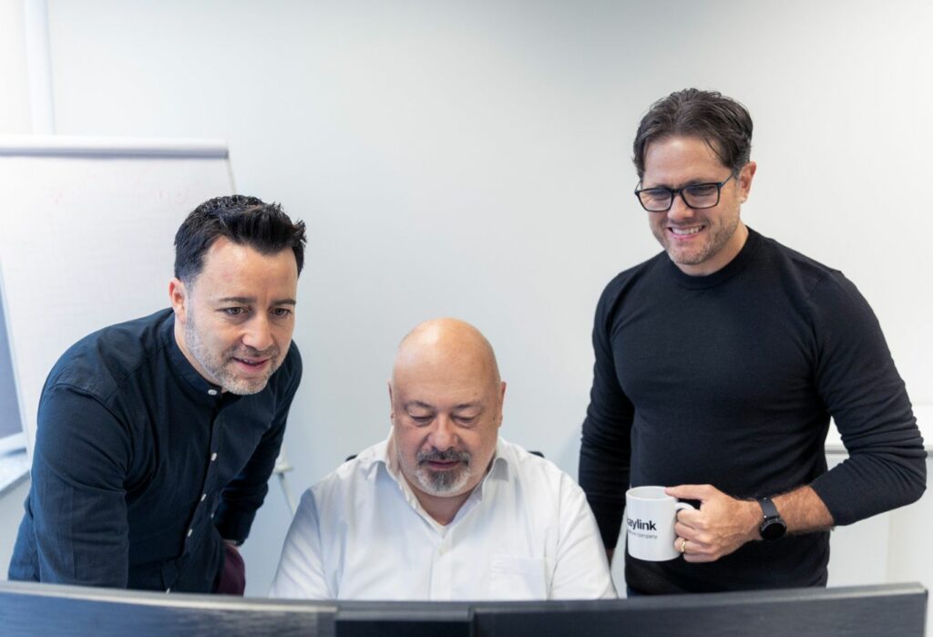 Drei Männer in einem Büro: Ein Mann in dunklem Hemd links, ein Mann in weißem Hemd in der Mitte vor einem Monitor, und ein Mann in schwarzem Pullover rechts mit einer Tasse in der Hand