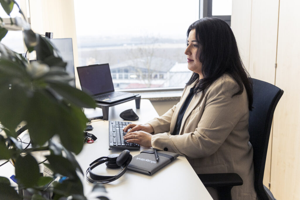 Frau sitzt an einem Schreibtisch in einem Büro mit Blick auf die Stadt, arbeitet an einer Tastatur, auf dem Tisch liegen ein Laptop, Kopfhörer und eine Pflanze