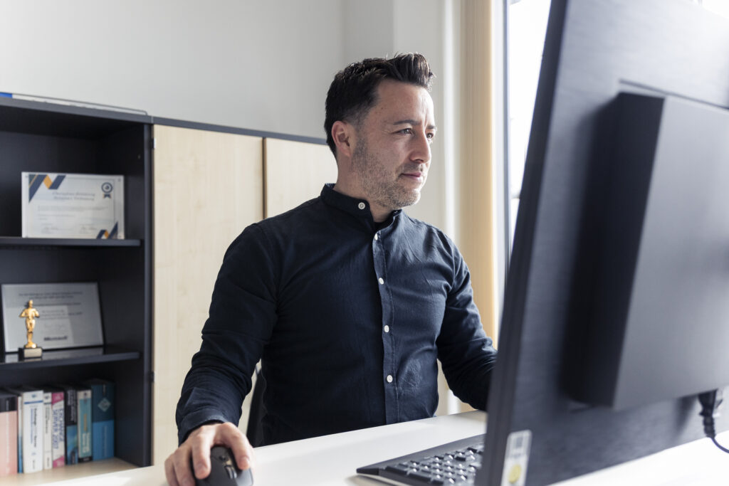 Mann in dunklem Hemd sitzt vor einem Computer im Büro mit Bücherregal und Auszeichnungen im Hintergrund