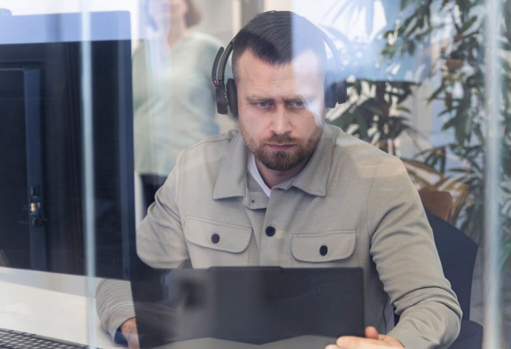 Mann mit Kopfhörern sitzt an einem Schreibtisch vor einem Laptop und arbeitet konzentriert in einem Büro mit Pflanzen im Hintergrund